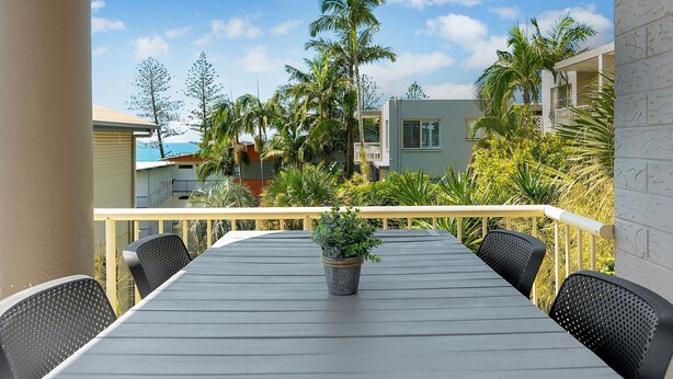 Un balcon avec une table et des chaises, entouré de plantes tropicales et offrant une vue sur les bâtiments et l’océan.