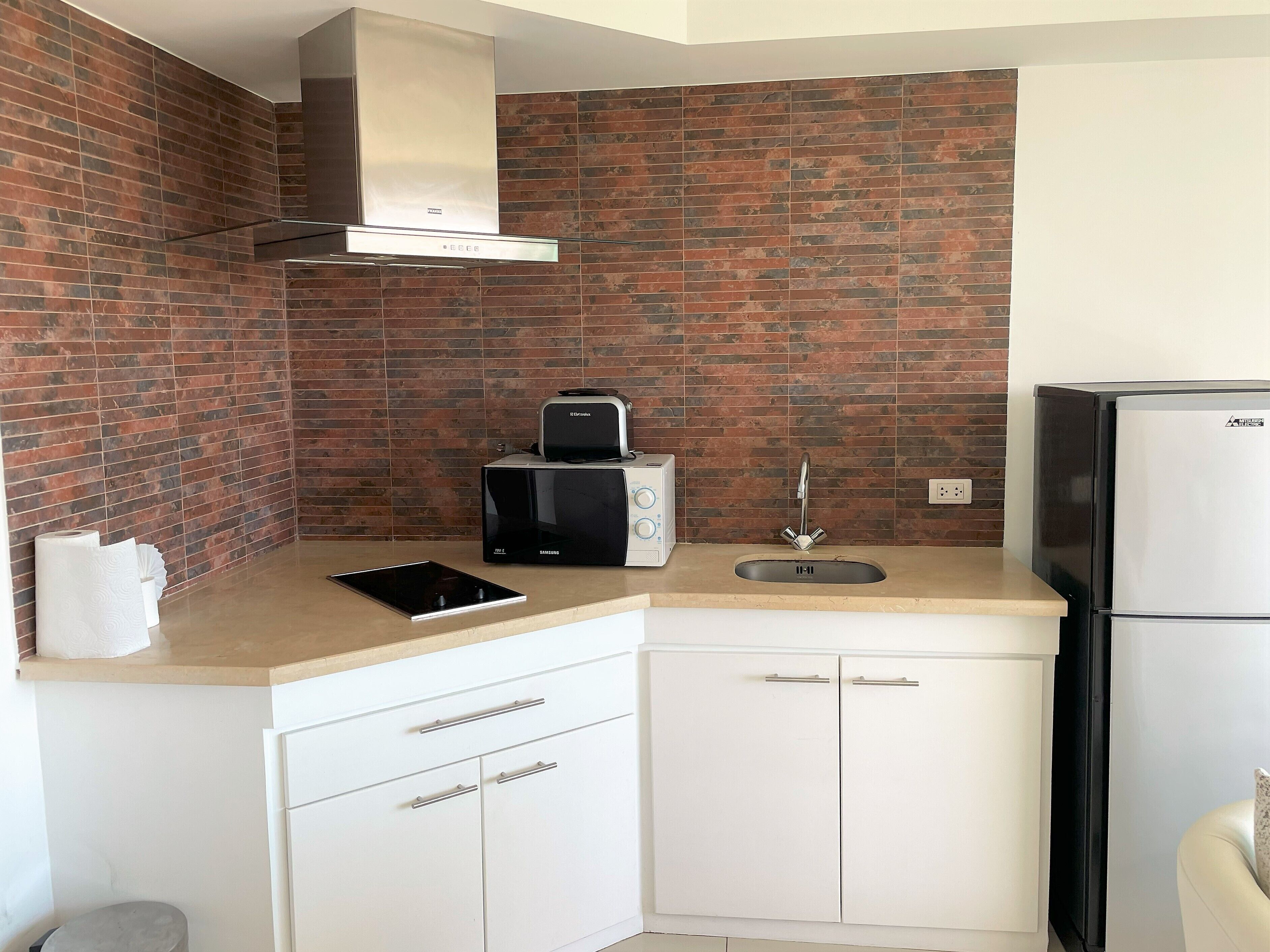 A compact kitchen with a brick backsplash, a stainless steel range hood, a black toaster, a sink, and a white refrigerator.