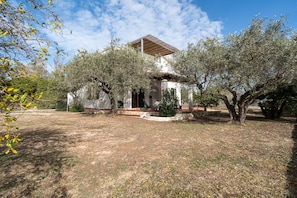 Una casa con porche cubierto, rodeada de olivos y un patio cercado.