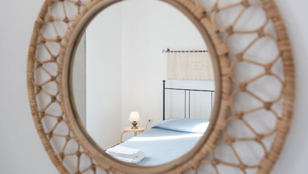 Un espejo redondo con un marco tejido, que refleja una cama con colcha azul y una lámpara blanca sobre una mesita de noche.