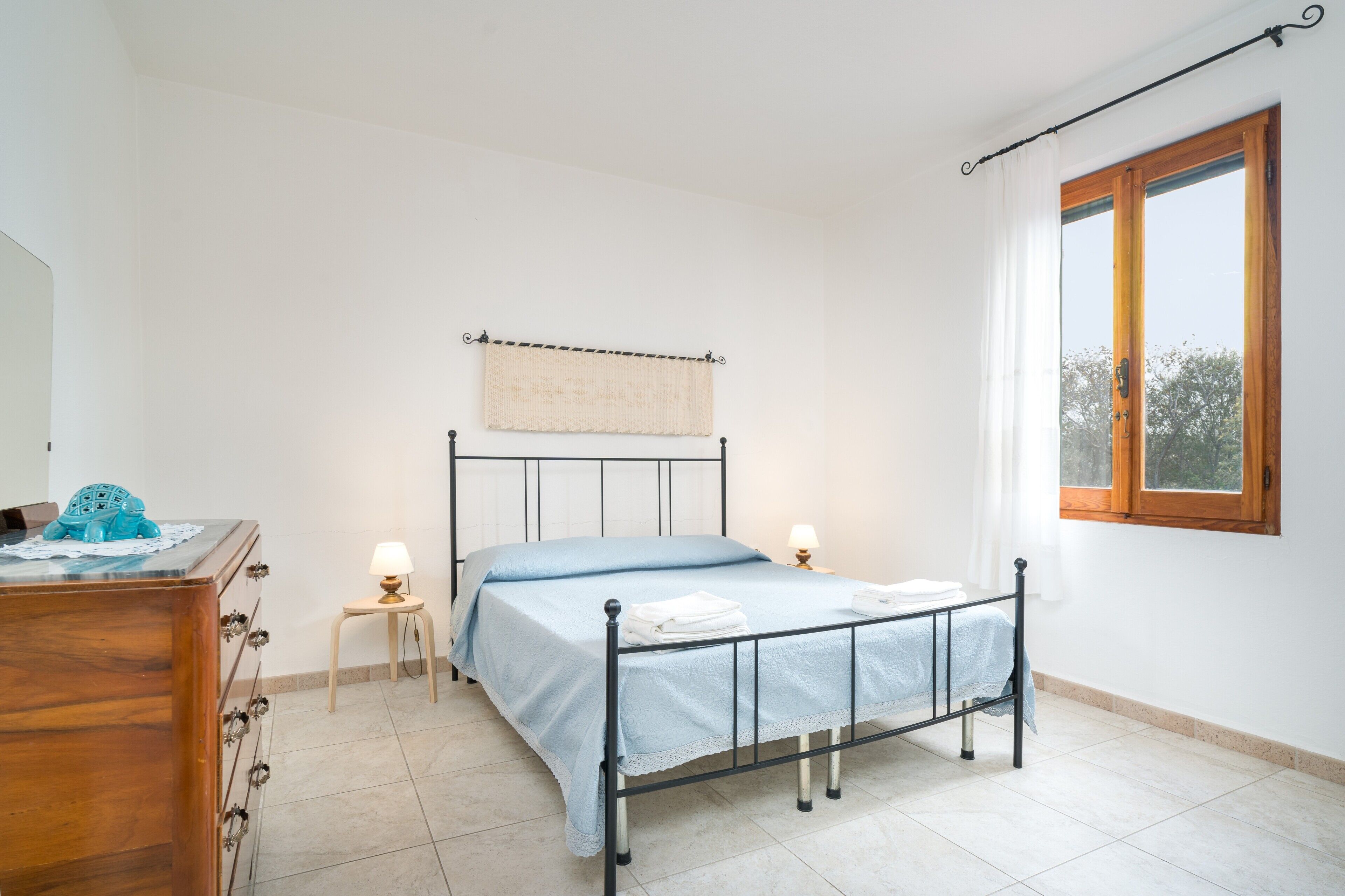 A bedroom with a bed, bedside lamps, a wooden dresser, a window with curtains, and a towel rack.