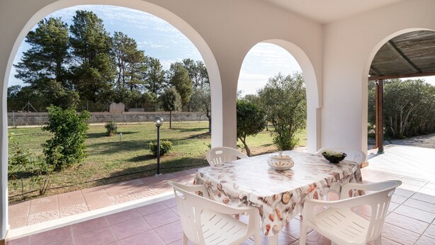 Un patio al aire libre con una mesa y sillas, vistas a un área verde con árboles, y un muro de piedra al fondo.