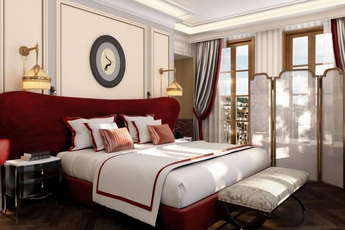 A spacious bedroom with a large bed, bedside tables, a bench, and a view of the city.