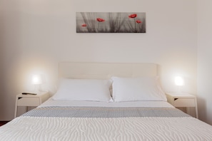 Un lit bien fait, avec du linge de lit blanc, deux tables de chevet avec des lampes et un tableau représentant des fleurs rouges accroché au mur.