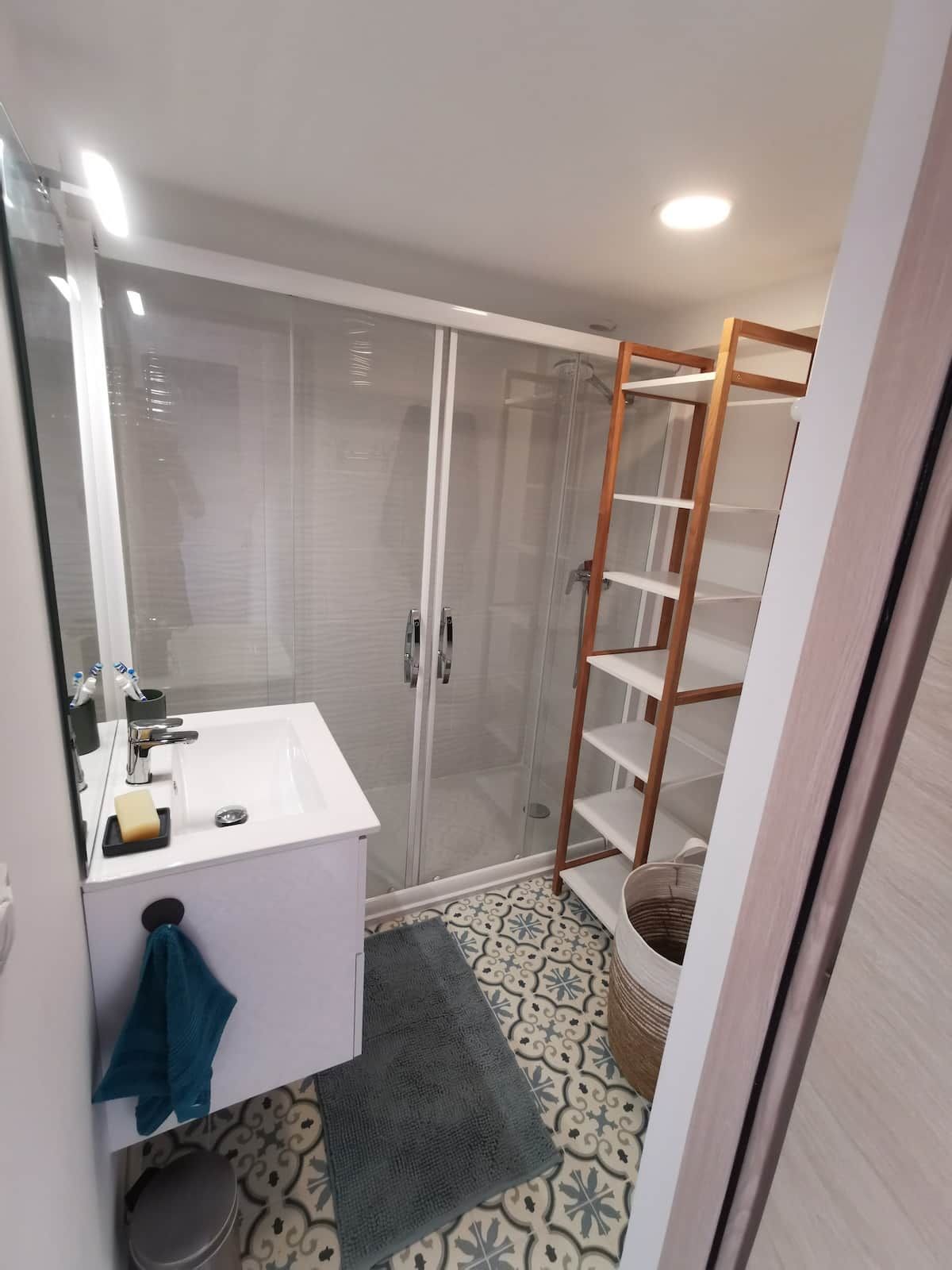 Une salle de bain avec un lavabo blanc, une cabine de douche en verre et une étagère en bois.
