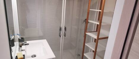 Une salle de bain avec un lavabo blanc, une cabine de douche en verre et une étagère en bois.