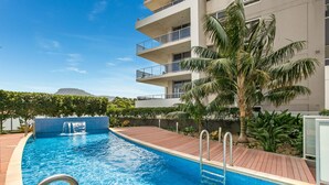 Una piscina con cubierta de madera, rodeada de palmeras y un edificio de varios pisos con balcones.