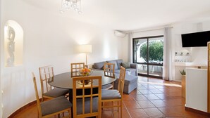Un coin repas avec une table ronde et des chaises, un canapé, un téléviseur et une fenêtre donnant sur un paysage verdoyant.