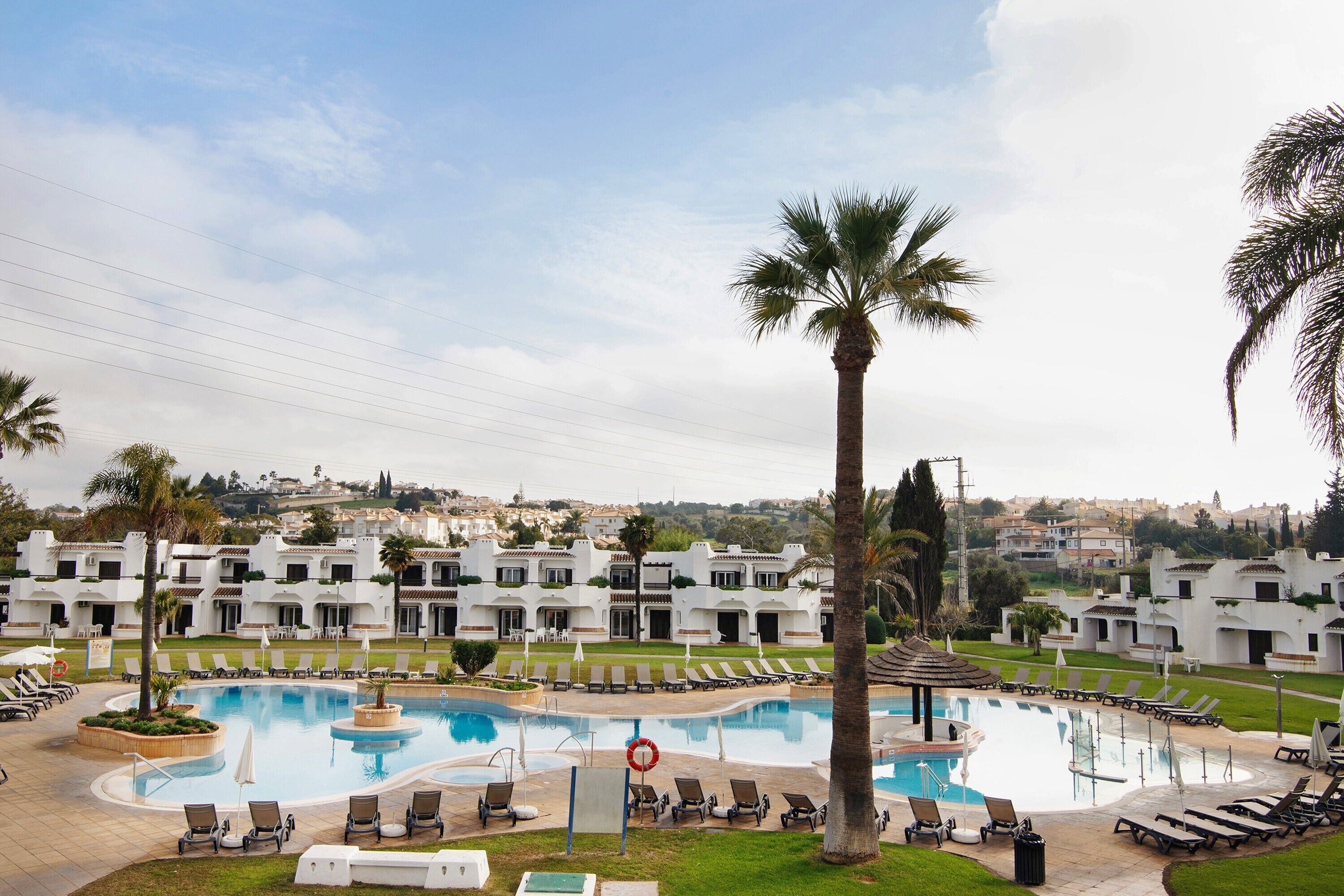 Un espace piscine équipé de chaises longues, d’une bouée de sauvetage et offrant une vue sur un quartier résidentiel avec des bâtiments blancs.