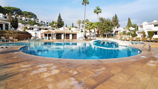 Une piscine avec des chaises longues, un restaurant et des bâtiments blancs entourés d’arbres.