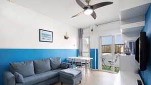 Una sala moderna con sofá, reposapiés y una mesa de comedor con sillas. Hay un ventilador de techo, un televisor montado en la pared y una ventana con vistas al exterior.