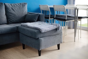 Un divano grigio con un poggiapiedi abbinato, una coperta blu in tessuto intrecciato e un'area da pranzo con sedie bianche e un tavolo.
