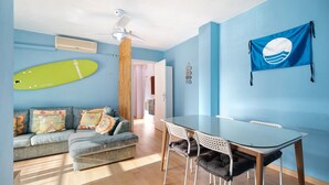 Une pièce avec un mur bleu, un ventilateur de plafond, une table à manger avec des chaises, un canapé avec des coussins et un élément de décoration représentant une planche de surf.