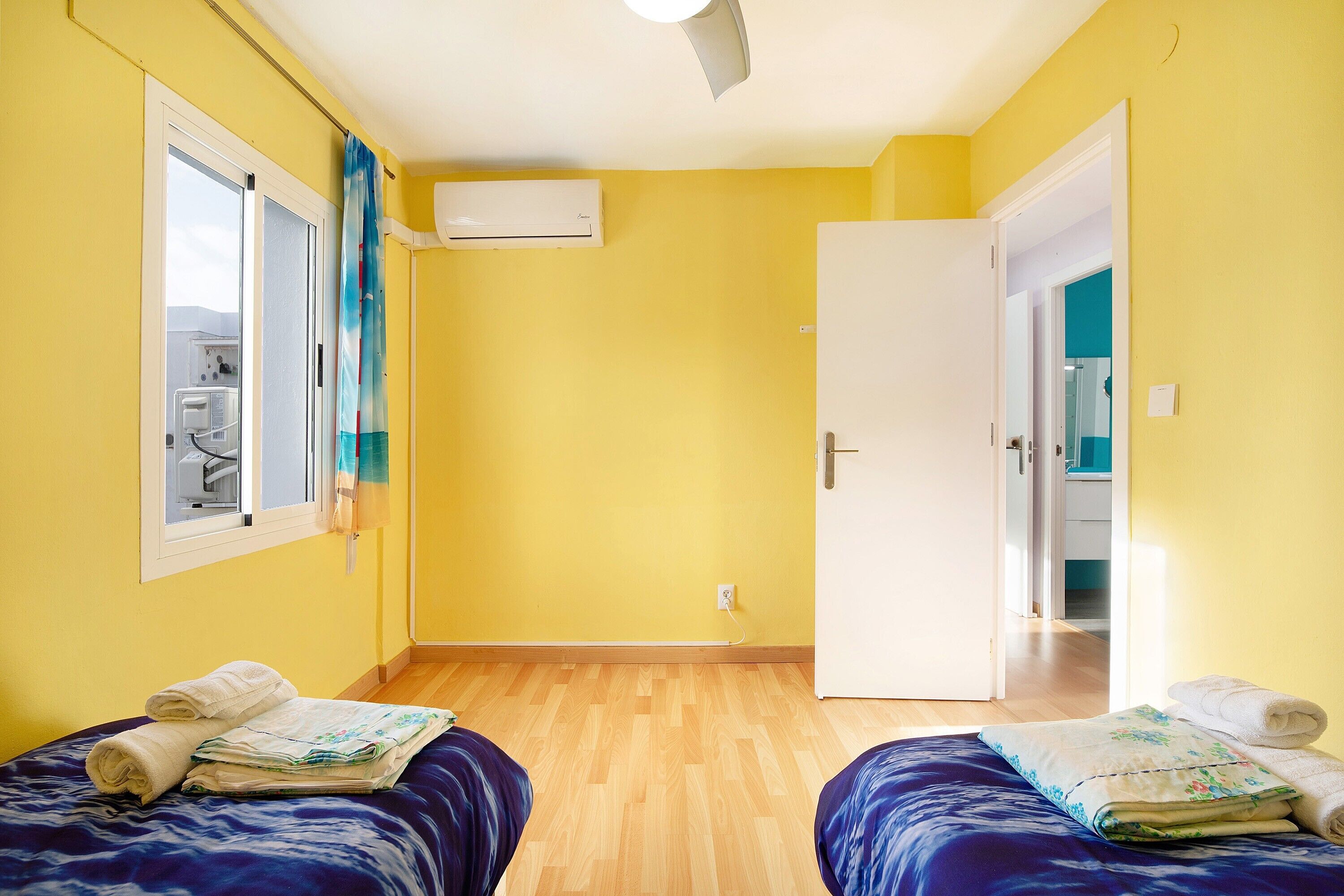 Une chambre avec deux lits, une fenêtre, un appareil de climatisation et une porte donnant sur une autre chambre.