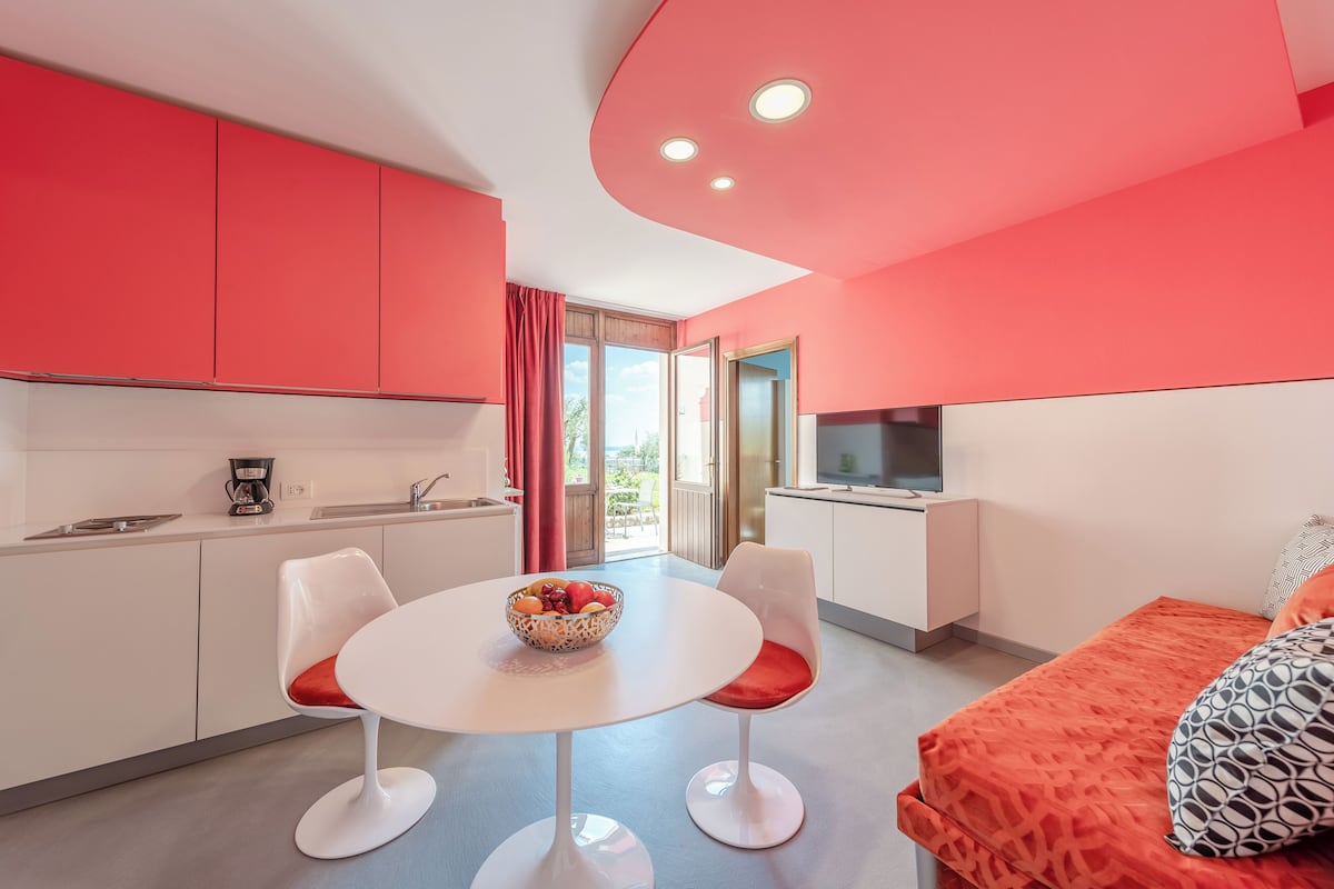 A modern kitchen with white cabinets and a red accent wall, a dining area with a white table and two chairs, and a bed with an orange cover.