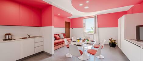 A modern kitchen with white cabinets and a red accent wall, a dining area with white chairs and a table, and a sofa in the background.