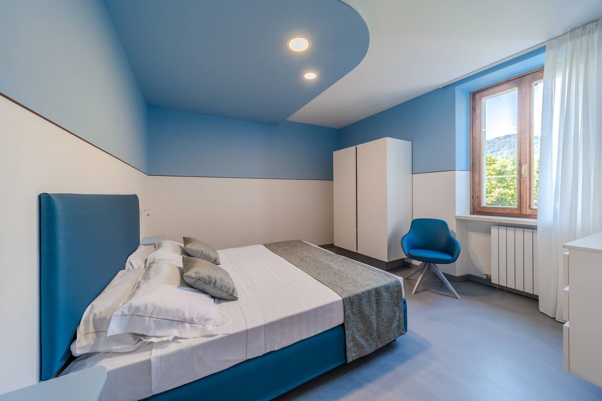 A bedroom with a bed, a chair, a wardrobe, and a window with curtains.