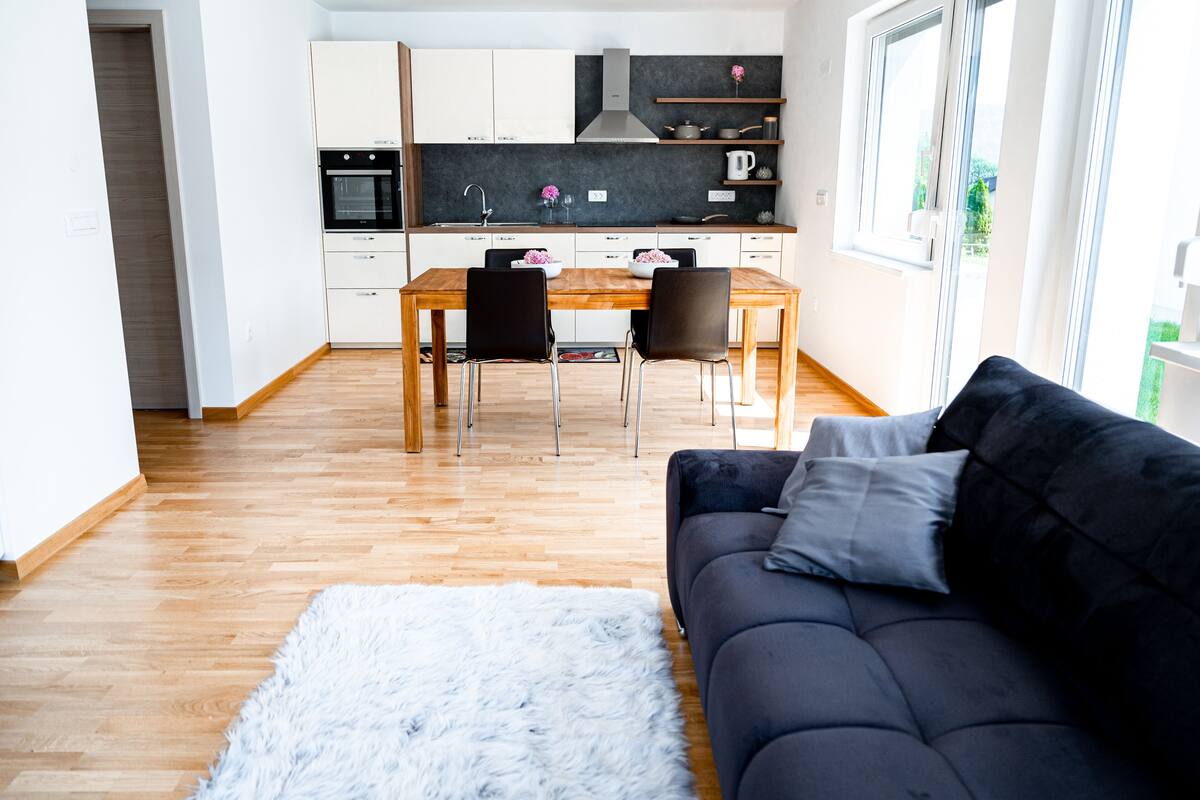 A modern kitchen with white cabinets, a dining table, and chairs. There is a black sofa with a fluffy rug in the living area.