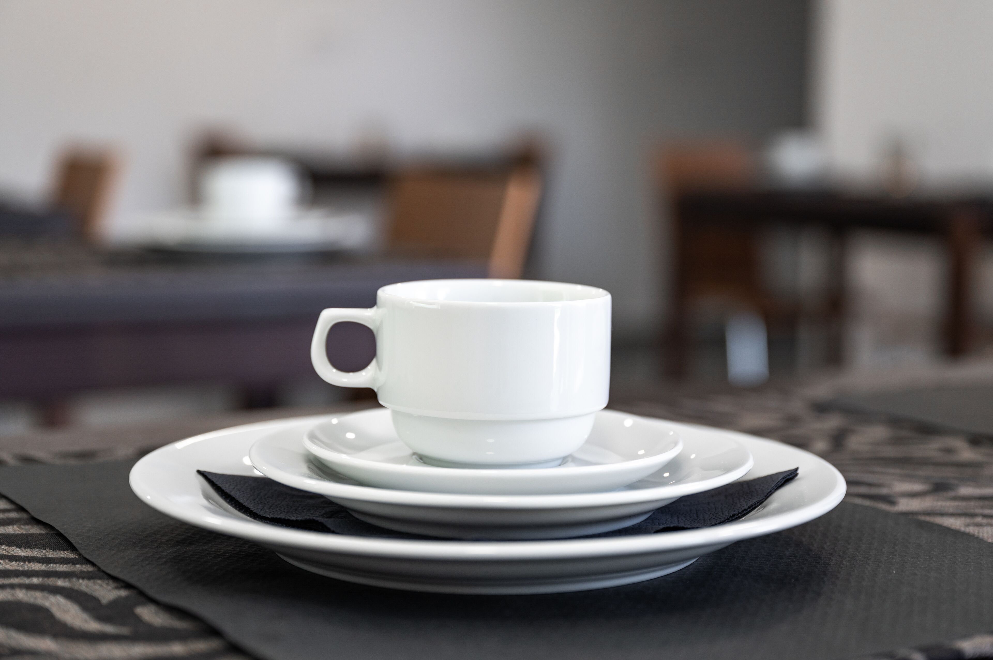 Una taza blanca sobre un platito y un plato, todo dispuesto sobre una mesa con un camino de mesa estampado.