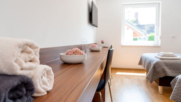 A modern hotel room with a wooden dining table, a bowl of pink flowers, neatly folded towels, a bed with a gray blanket, and a window with a view of buildings.