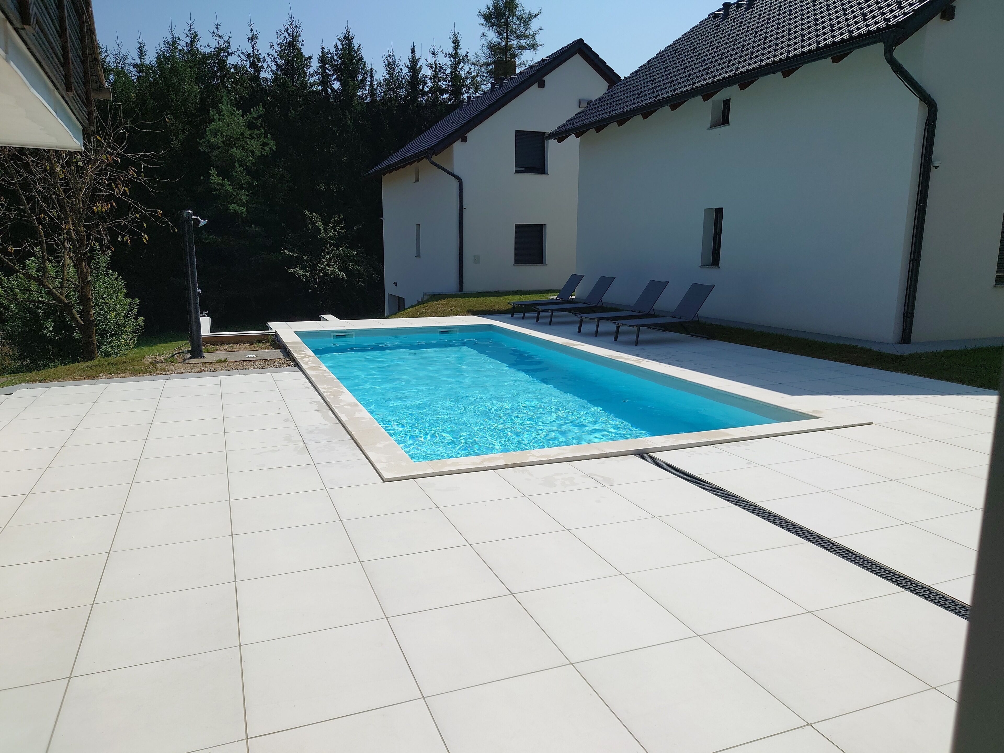 A rectangular swimming pool with a tiled deck, surrounded by a white building with a tiled roof and a grassy area with lounge chairs.