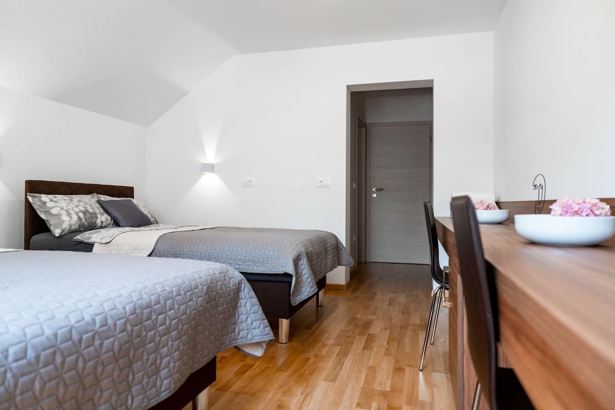 A bedroom with two beds, a wooden floor, a desk with a bowl of flowers, and a door leading to another room.