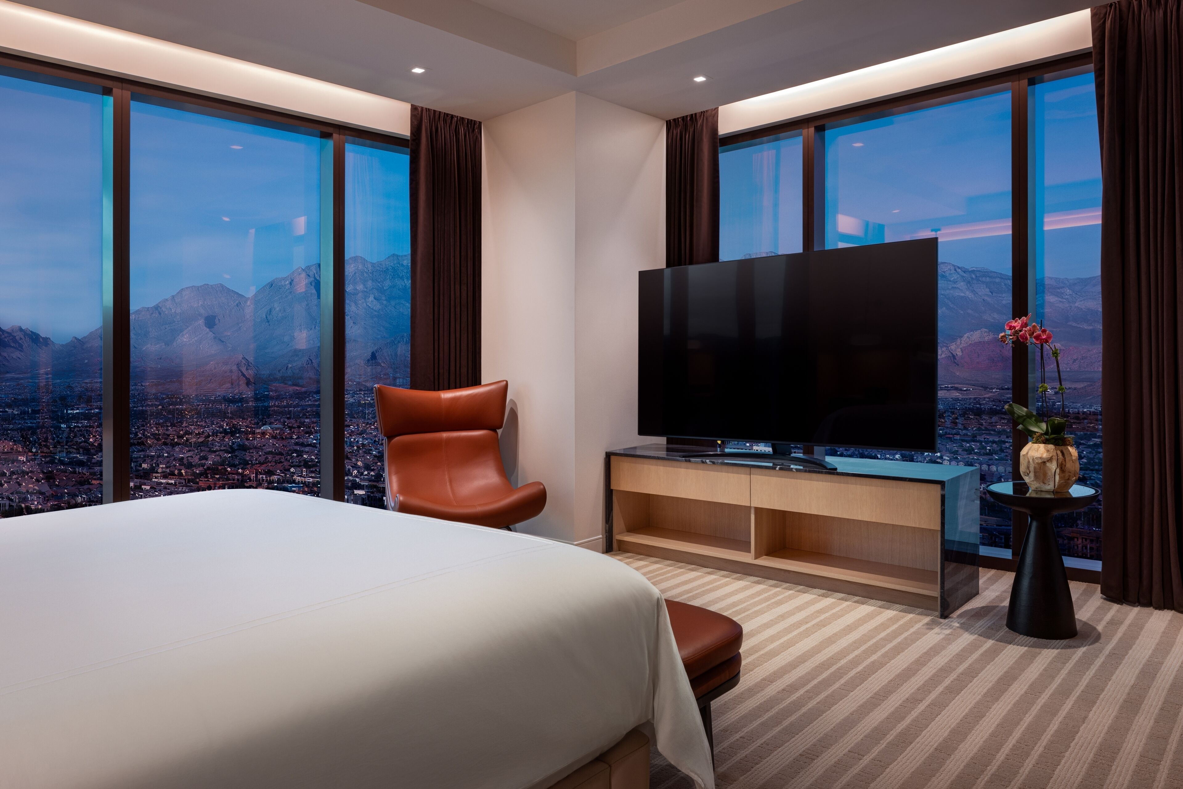A hotel room with a large bed, a flat-screen TV, a leather chair, a small table with a vase, and a city view through large windows.