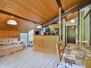 A cozy cabin interior with a wooden ceiling, a sofa with patterned cushions, a dining table, and a kitchen area with a microwave and cabinets.
