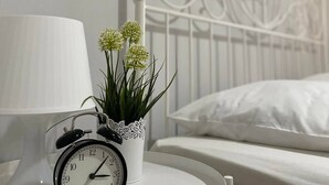 Un lit bien fait, avec une literie blanche, une table de chevet blanche ornée d’une horloge et d’une petite plante en pot, et une tête de lit en métal blanche.