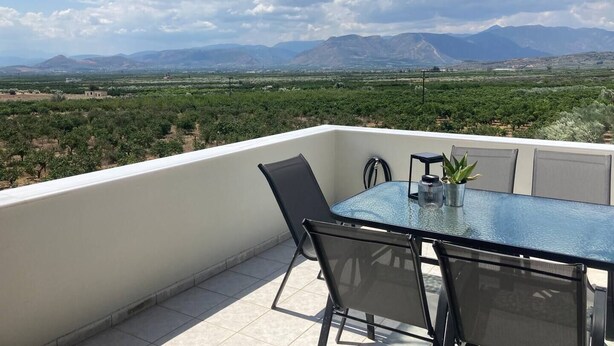 Un balcon doté d’une table et de chaises en verre, offrant une vue sur un vignoble et des montagnes lointaines.