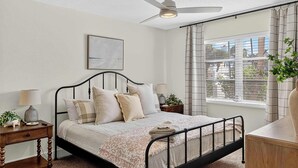 A bedroom with a bed, bedside tables, a ceiling fan, a window with curtains, and a view of buildings outside.