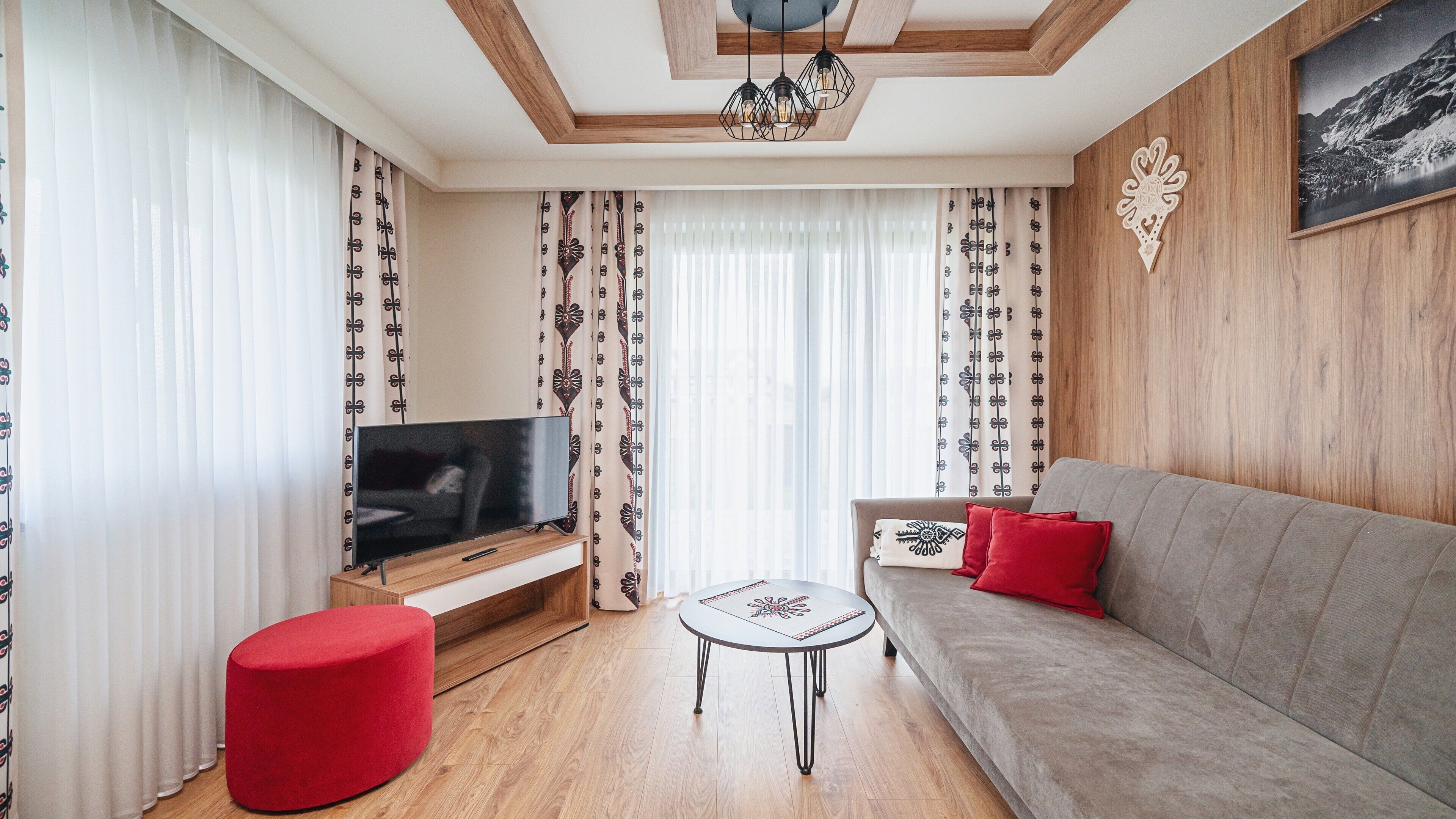 Una sala de estar moderna con un televisor de pantalla plana sobre un soporte de madera, un sofá gris con cojines rojos, un reposapiés rojo y cortinas estampadas.