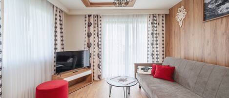 Una sala de estar moderna con un televisor de pantalla plana sobre un soporte de madera, un sofá gris con cojines rojos, un reposapiés rojo y cortinas estampadas.