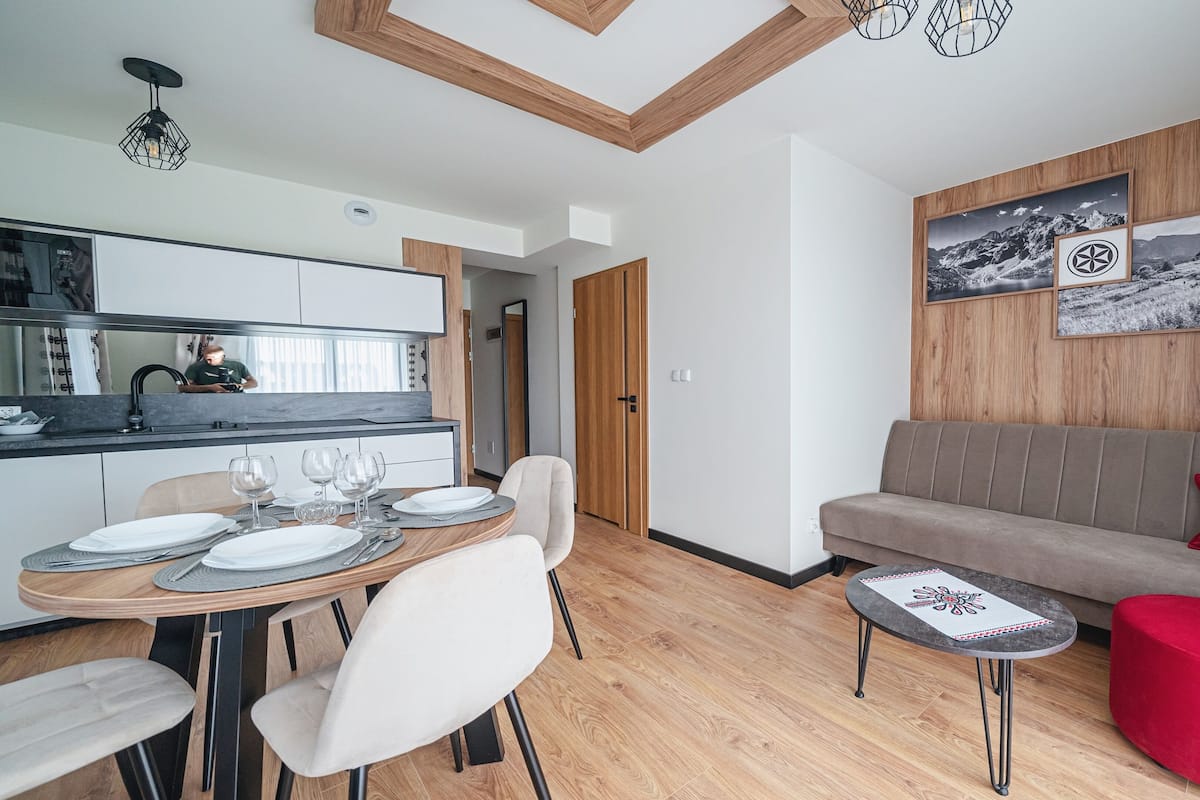 Un salon moderne comprenant un canapé, une table à manger dressée avec des assiettes et des verres, un pouf rouge et une petite table sur laquelle est posé un objet décoratif.