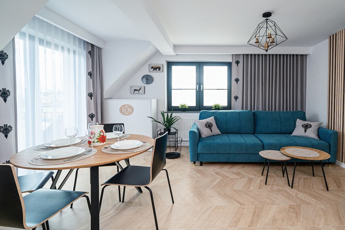 Un salon moderne avec un canapé bleu, une table à manger dressée pour deux personnes et un lustre aux motifs géométriques.