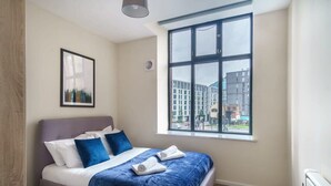 A modern bedroom with a large window, a bed with blue bedding, and a framed picture on the wall. - Bright 2 Bedroom Apartment in Central Manchester (Manchester)