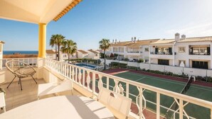 Un balcon donnant sur un court de tennis et une piscine, avec des bâtiments blancs en arrière-plan et un ciel dégagé.