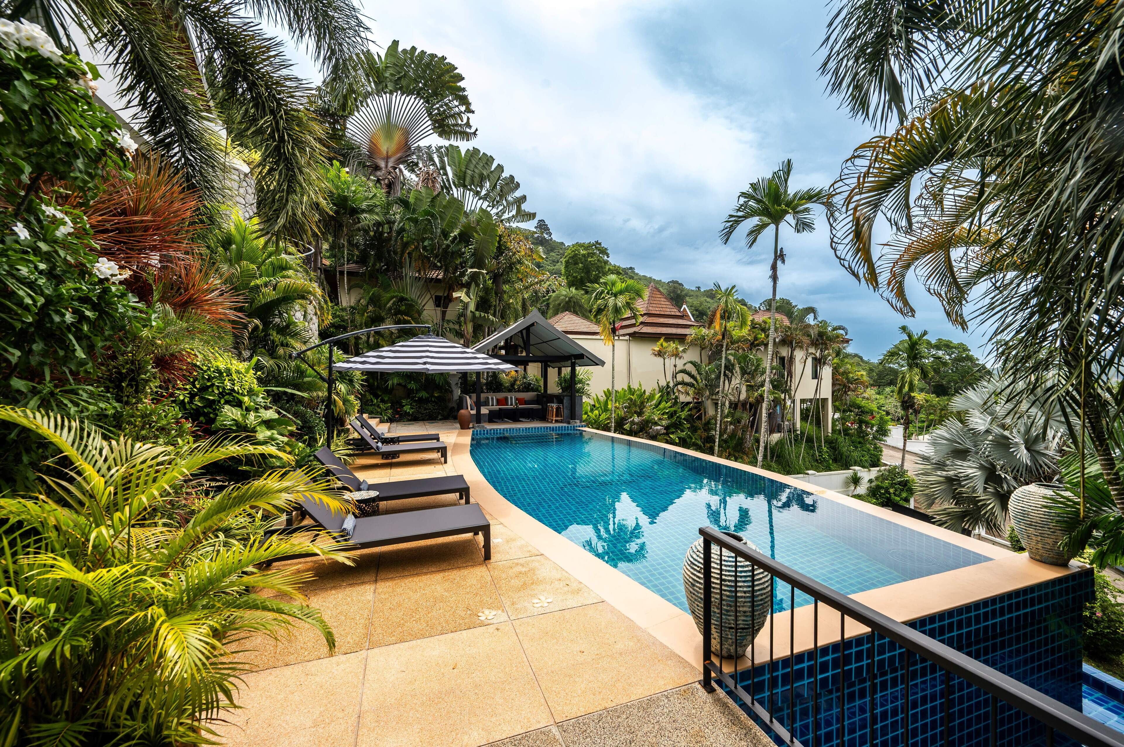 Une piscine entourée d’une végétation tropicale luxuriante et de palmiers, avec des chaises longues et un espace salon abrité.
