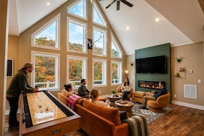 A spacious living room with a large wooden table, comfortable seating, a fireplace, and large windows offering a view of autumn foliage.