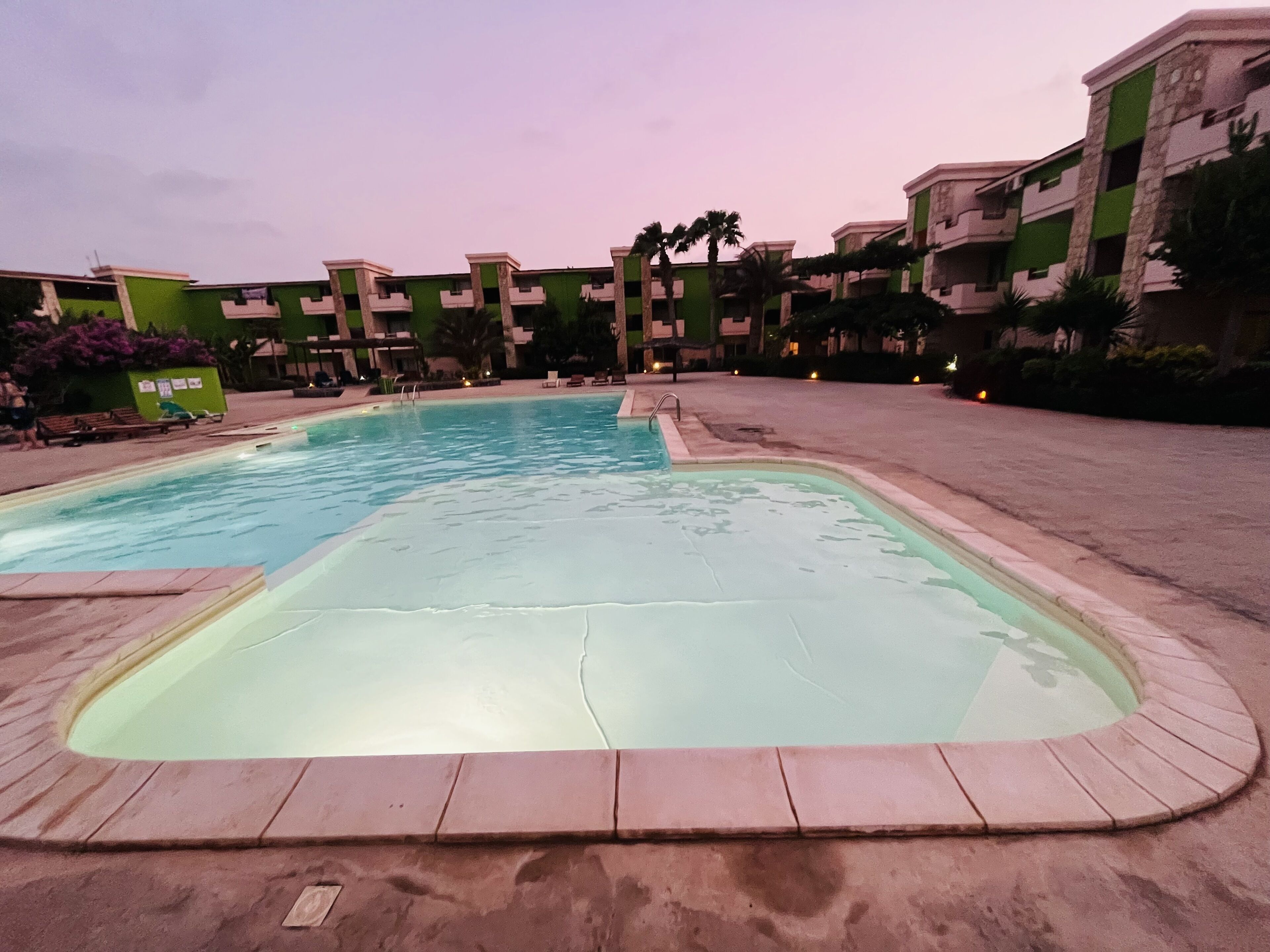 A swimming pool surrounded by multi-story buildings with balconies, a clear sky, and some greenery.