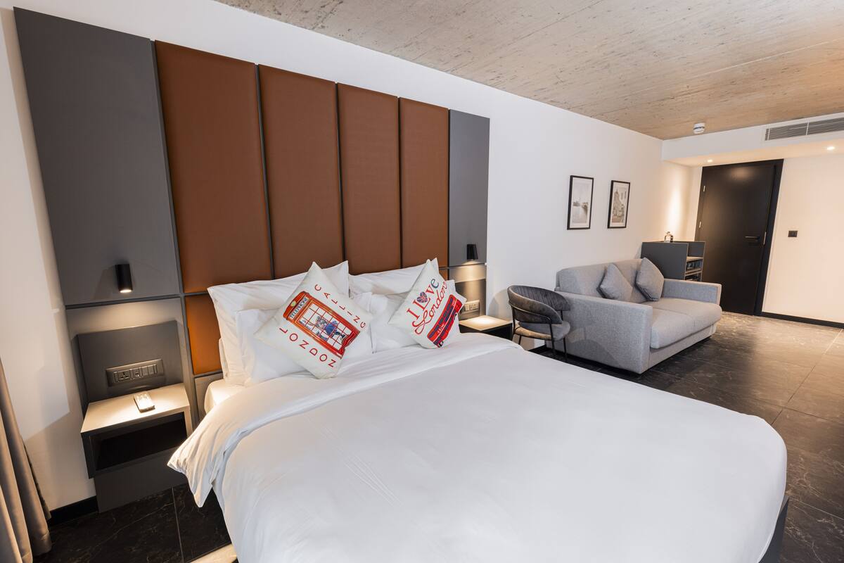 Habitació d'hotel moderna amb un llit gran, un sofà gris i una taula amb una làmpada.