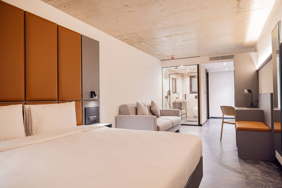 Habitació d'hotel moderna amb un llit gran, un sofà, una taula de treball i una cadira. Es veu un bany per darrere d'una porta de vidre.