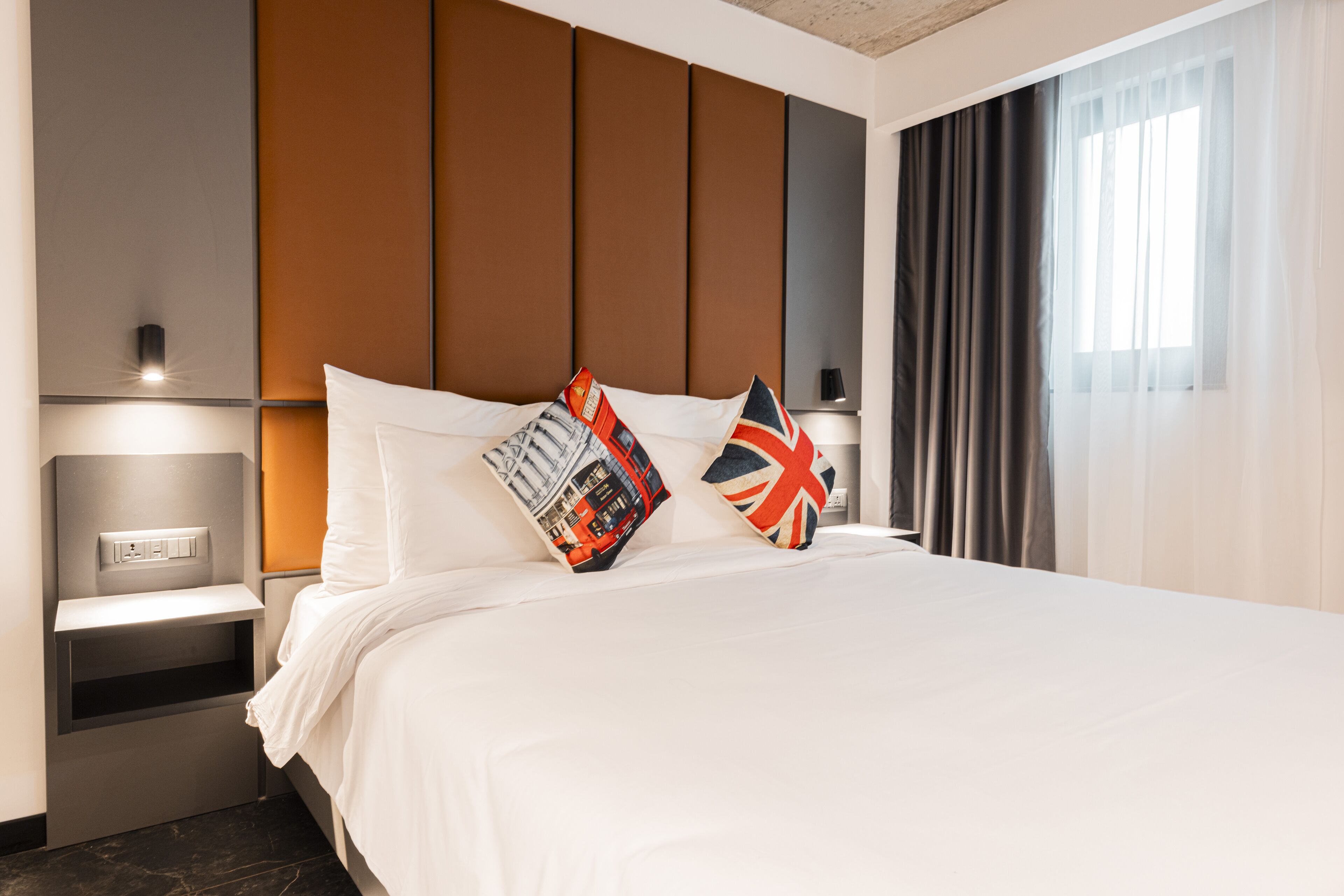 A modern hotel room with a large bed, two pillows with British flags, a nightstand with a lamp, and a window with curtains.