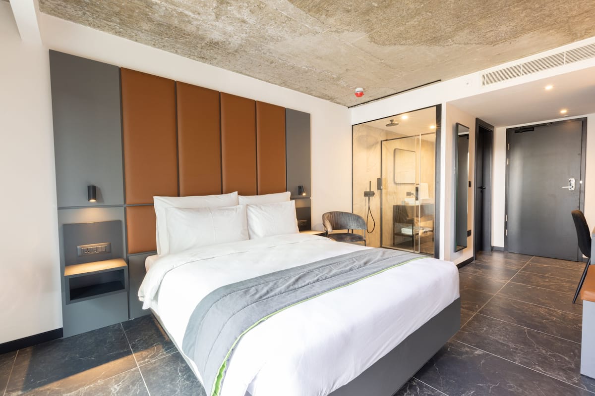 Habitació d'hotel moderna amb un llit gran, un armari encastrat, una taula de treball i un bany visible a través d'una porta de vidre.