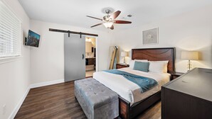 Une chambre d’hôtel comprenant un lit, un ventilateur de plafond, une table de chevet, une lampe, un banc et une porte donnant accès à une salle de bain.