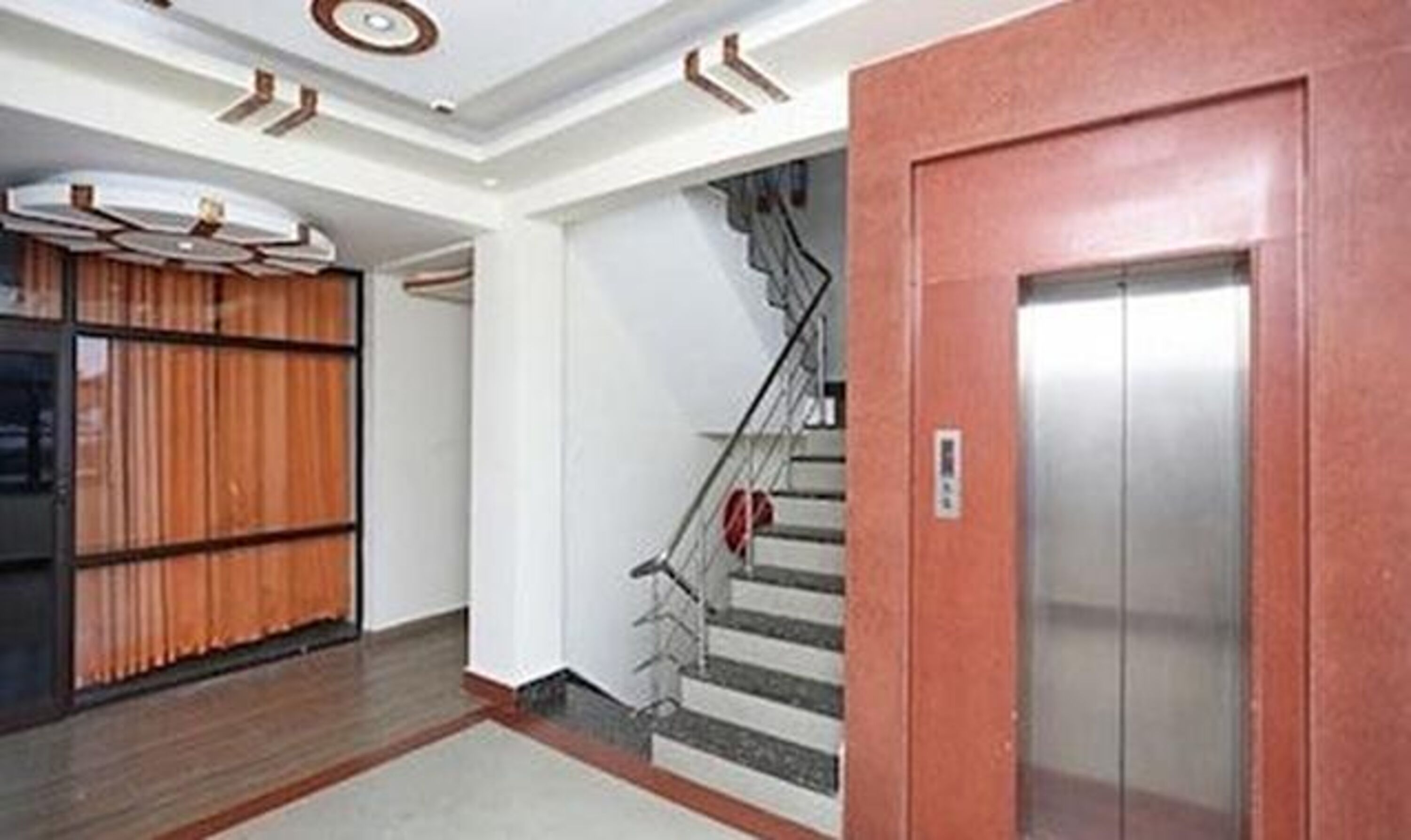 A hotel lobby with a wooden reception desk, a staircase with a red handrail, and an elevator.