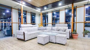 A modern lounge area with a sectional sofa and ottomans, large windows, and a wooden floor. - Fabhotel Wildflower Inn (Nainital)