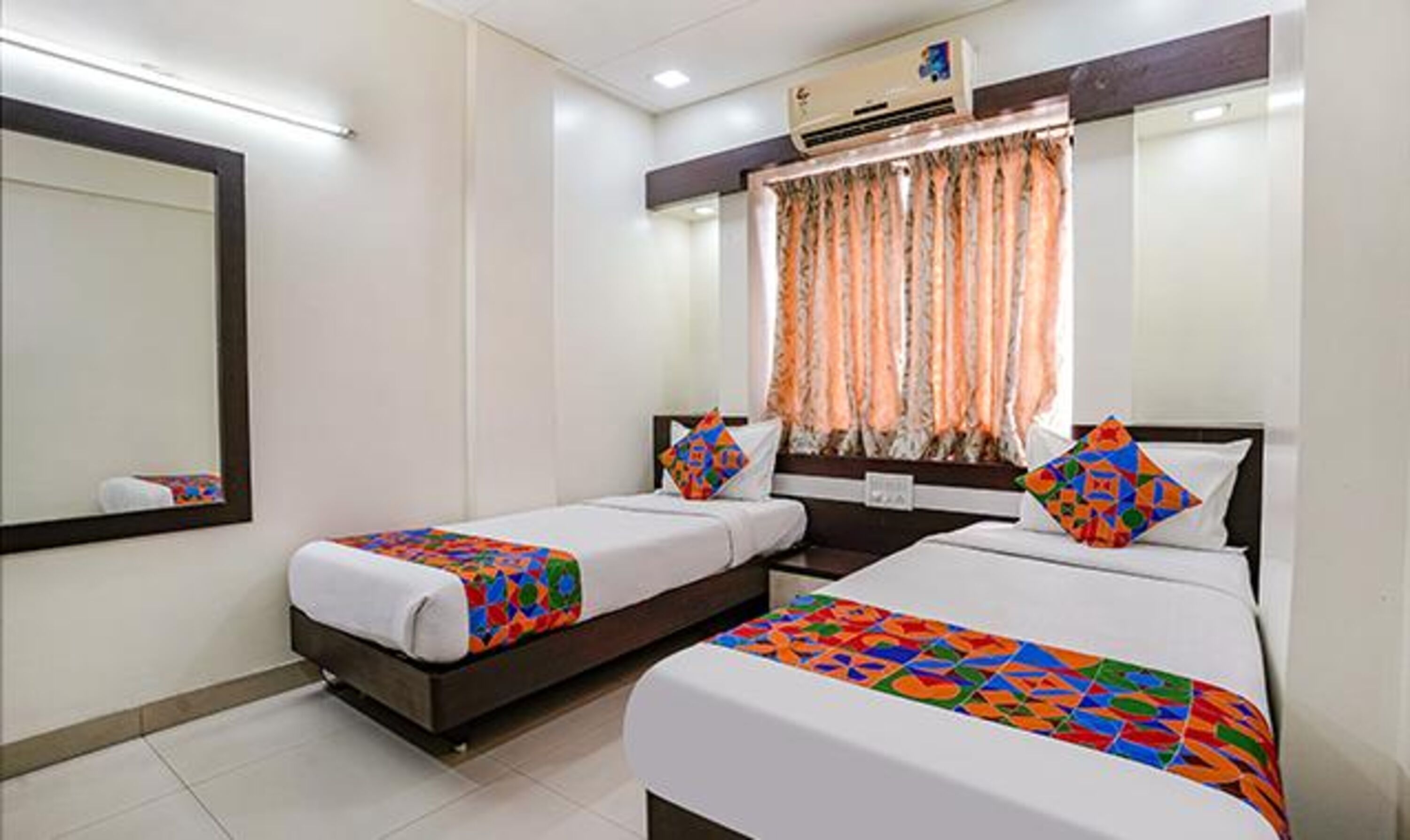 Una habitación de hotel con dos camas, un espejo grande, un equipo de aire acondicionado y una ventana con cortinas.