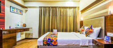 Quarto de hotel com cama, mesa de cabeceira, escrivaninha e cortinas.