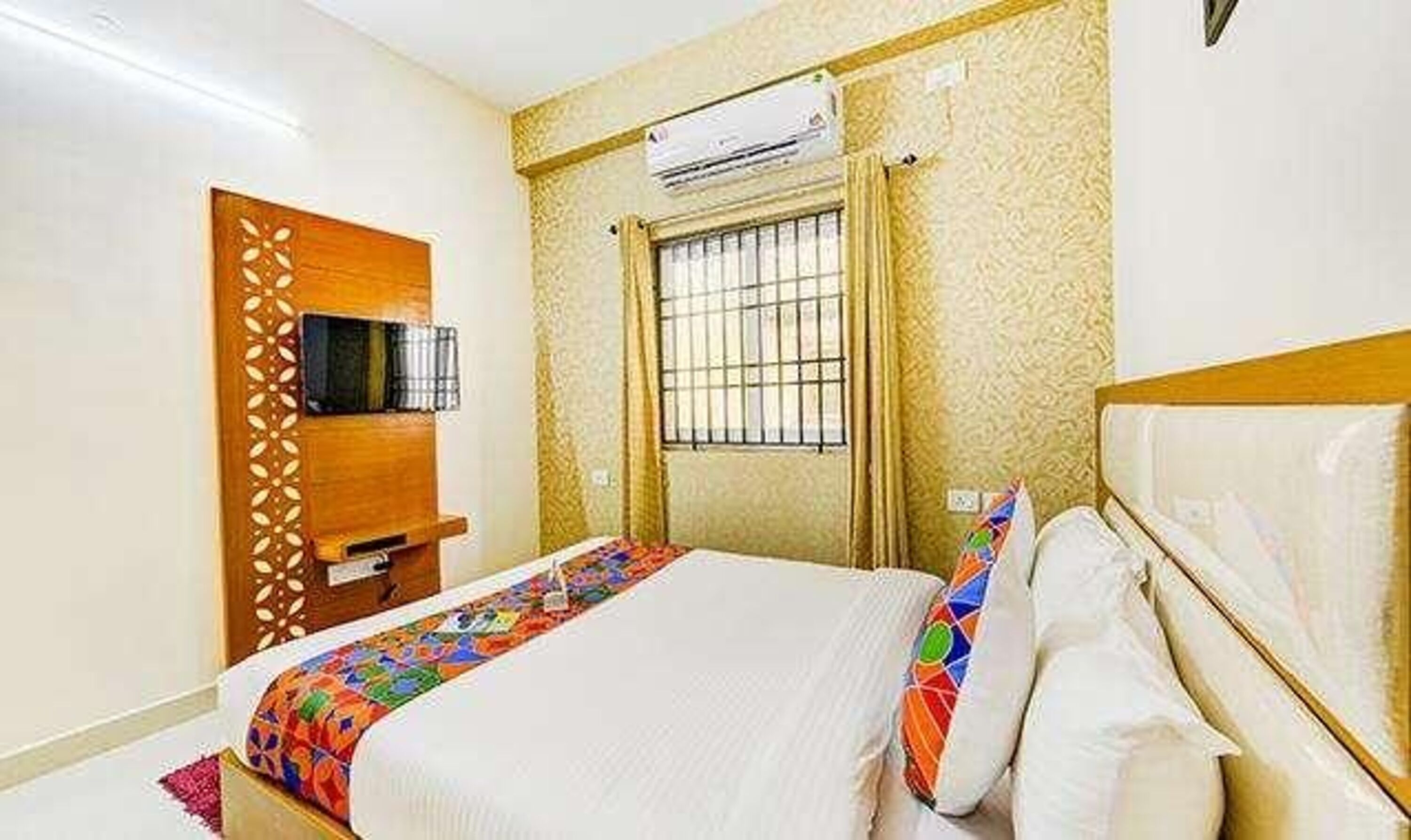 A hotel room with a bed, a TV on a wooden stand, an air conditioning unit, and a window with bars.